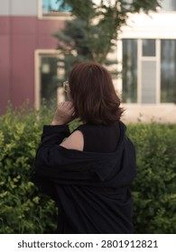 A vertical, eye-level medium shot from behind of a mid-adult woman with shoulder-length, thick auburn-brown hair. The woman is captured from a rear three-quarter view, looking away from the camera toward a building in the background. Her right hand is raised slightly toward her face, and the edge of clear-framed eyeglasses is visible on her profile. She wears a casual black sleeveless top underneath an oversized black button-up shirt that is styled loosely off her right shoulder. She stands in front of a dense row of manicured green garden shrubs. The background features a modern residential or office building with large glass windows and a mauve-colored facade, softly out of focus due to a shallow depth of field in warm, natural evening light.