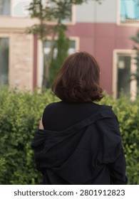 A vertical, eye-level medium shot from behind of a mid-adult woman with shoulder-length, thick auburn-brown hair. The woman is captured from a rear three-quarter view, looking away from the camera toward a building in the background. Her right hand is raised slightly toward her face, and the edge of clear-framed eyeglasses is visible on her profile. She wears a casual black sleeveless top underneath an oversized black button-up shirt that is styled loosely off her right shoulder. She stands in front of a dense row of manicured green garden shrubs. The background features a modern residential or office building with large glass windows and a mauve-colored facade, softly out of focus due to a shallow depth of field in warm, natural evening light.