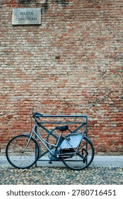 Vertical, eye-level shot capturing a classic silver ladies' city cruiser bicycle parked securely against a weathered red brick wall in Italy. The vintage bike features a black leather saddle seat, matching black handlebars, a rear fender splash guard, and a rear wheel lock device. It is parked parallel to a large, textured masonry facade composed of multi-toned orange and red clay bricks. High up on the wall, an authentic marble Italian street plaque marks the location as 