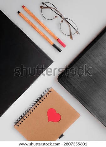 A vertical flat lay composition showing a black folder, a laptop lid corner, two thin fineliner pens, glasses, and a brown spiral notebook with a red heart on a white background.