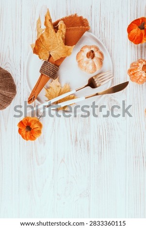 Vertical flat lay of fall table setting with orange napkin, ceramic pumpkin, cutlery and maple leaves on rustic white wood