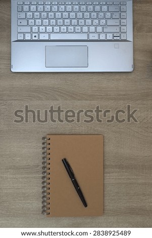 Vertical format flat lay composition showing the lower half of a laptop keyboard, a spiral notepad, and a black pen on a rustic wooden desk.