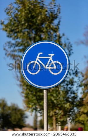 Vertical frame of Dutch traffic sign depicting a white bicycle on a blue round metal board meaning an obligated bike path for bikeriders against a blue sky with out of focus trees in the background