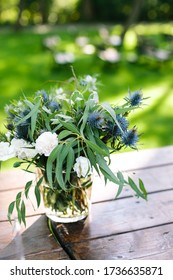 Vertical Frame. Focus On The Wedding Flower Centerpiece Made From Cloves, Carnation, Eucalyptus, Thistle In A Transparent Glass Vase On A Wooden Tabletop Against A Green Lawn.