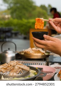 A vertical frame of a handheld grilled bite above a barbecue hot pot, with shallow focus and outdoor dining context. The frame is suited for Asian restaurant, Thai barbecue hot pot, shared dining, grilled pork, seafood hot pot, cooking process, and casual meal concepts.