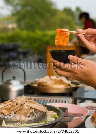 A vertical frame of a handheld grilled bite above a barbecue hot pot, with shallow focus and outdoor dining context. The frame is suited for Asian restaurant, Thai barbecue hot pot, shared dining, grilled pork, seafood hot pot, cooking process, and casual meal concepts.