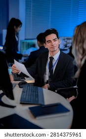 Vertical frame of a young developer with documents beside his blonde teammate initialing the reviewed sheet, in a dark blue technology office lit by screens with quiet focus.
