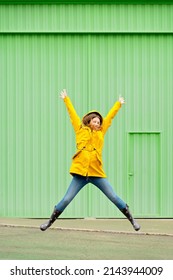 Vertical front view of woman jumping in a yellow raincoat on the street. Full length body of woman under the rain isolated leaping on green wall. People isolated in background concept. - Shutterstock ID 2143944009