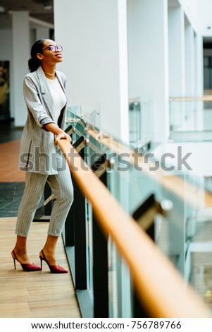 Vertical full height of elegant african woman in glasses and gray suit standing at railing in business center or shopping mall. Side view. Looking up. Lifestyle, leisure and people concept.