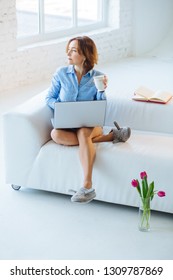 Vertical Full Height Of Mature Woman In Blue Shirt And Slippers Sits On Couch Feeling Harmony Excited, Working At Home With Laptop In Comfort Atmosphere Break Resting In White Wall Background.