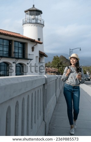 vertical full length of asian Japanese lady tourist admiring beauty of sunset while strolling on stone bridge with a lighthouse building at background in santa Barbara usa