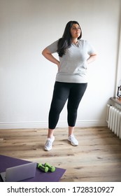 Vertical Full Length Image Of Overweight Young Woman Wearing Sports Clothes And Sneakers Doing Cardio Workout At Home. Plus Size Female Training Indoors Using Laptop, Dumbbells And Fitness Mat