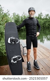 Vertical full length portrait of bearded senior man wearing wakeboarding gear and holding board smiling at camera standing on pier by lake outdoors