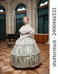 Vertical full length portrait of beautiful young woman wearing classic ballgown standing in spotlight in palace hall