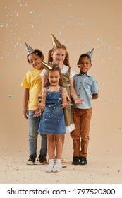 Vertical Full Length Portrait Of Diverse Group Of Children Wearing Party Hats Smiling At Camera While Standing Under Colorful Confetti Burst In Studio