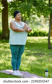 Vertical Full Length Portrait Of Mature Black Woman Enjoying Yoga Outdoors In Green Park And Standing With Eyes Closed Doing Breathing Exercises