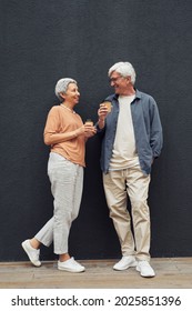 Vertical Full Length Portrait Of Modern Mature Couple Looking At Each Other And Smiling While Standing By Grey Wall