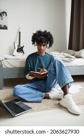 Vertical Full Length Portrait Of Teenage African-American Student Sitting On Floor At Home Or In College Dorm And Studying