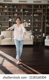 Vertical Full Length View Happy Relaxed Beautiful Young Hispanic Woman Looking At Camera, Making Funny Moves, Dancing Barefoot In Modern Living Room, Listening Favorite Music In Wireless Earphones.