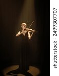 Vertical full shot of concentrated female violin player performing solo concerto under dramatic projector light in dark studio on black background, copy space