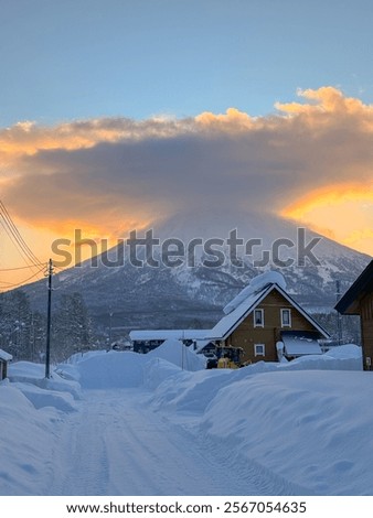 VERTICAL: Glowing orange sunset illuminates Mt. Yotei towering above the wintry wonderland near Niseko, Japan. Idyllic shot of a residential part of the snow-covered countryside near Niseko, Japan.