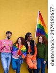 Vertical. Group of diverse LGBT young people leaning on wall yellow. Smiling friends celebrating gay pride day enjoying together and showing rainbow flags. Generation z relationships and copy space