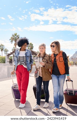 Vertical group mature diverse tourist women empowered friends walking with luggage while holding and looking travel map in their hands. Tourism people enjoying their vacation on the sunny city street