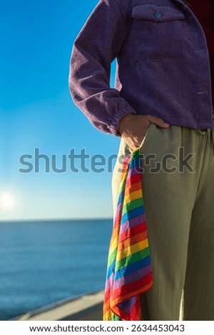 Vertical half-figure image captured from shoulder to knee. One hand rests on the pocket of the pants, from which a scarf with rainbow colors, symbol of LGTBI pride, is peeking out.
