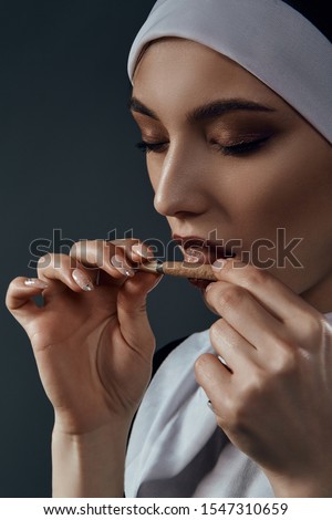 Vertical half-turn close-up portrait of a nun, posing on a black background. She wearing dark nun's clothing. The nun brings a cigarette to her mouth and licking it.  