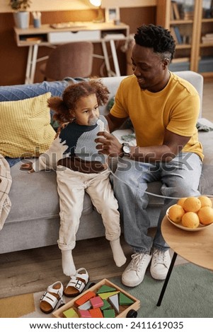 Vertical high angle shot of friendly Black father supporting his daughter during inhalation procedure holding nebulizer mask