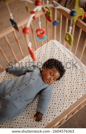 Vertical high angle shot of sleeping Black girl dressed in blue knitted jumpsuit peacefully lying on back in baby crib with calming musical mobiles, hanging above child in home setting
