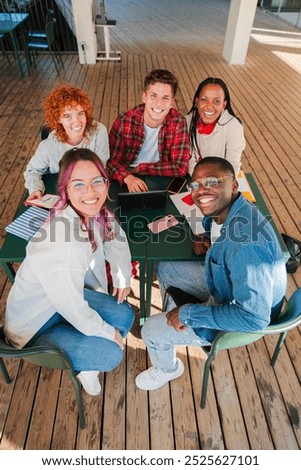 Vertical high angle view of diverse group of smiling students looking at camera, working together around a table, with notebooks and smartphones, studying and collaborating in a casual setting indoors