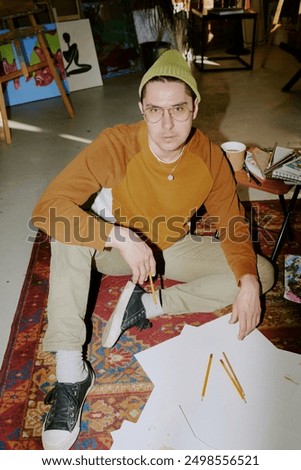 Vertical high angle view flash shot of young Caucasian male artist sitting on vintage carpet on floor in his studio