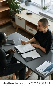 Vertical high angle view shot of cheerful young Caucasian woman sitting at desk in office having conversation with African American job applicant