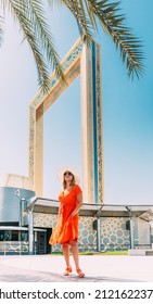 Vertical High quality photo. Young Caucasian Woman In Red Dress And In Summer Hat Standing Near Dubai Frame And Posing For Camera. View Of Dubai Frame In Zabeel Park On Sunny Blue Sky Under Palm