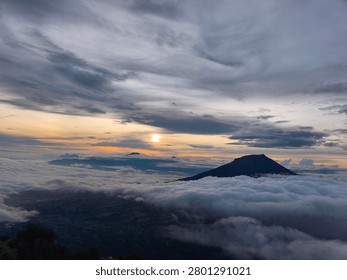 Vertical high-angle composition of a massive volcanic mountain silhouette rising above a dense sea of clouds, capturing a moody and dramatic twilight atmosphere with a subtle golden sunrise glow on the horizon under an overcast dark blue sky.