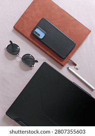 A vertical high-angle flat lay shot showcasing a modern smartphone, a brown leather notebook, stylish sunglasses, a pen, and a laptop arranged on a smooth pink surface