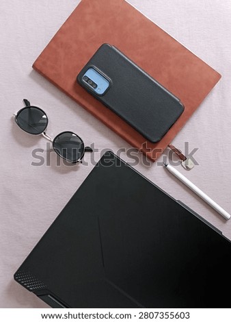 A vertical high-angle flat lay shot showcasing a modern smartphone, a brown leather notebook, stylish sunglasses, a pen, and a laptop arranged on a smooth pink surface