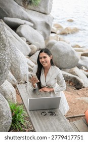 A vertical high-angle shot captures a smiling Thai woman looking off-camera while holding her smartphone in Phuket, Thailand. She is leaning against a wooden table with an open laptop, representing a modern, flexible workspace. The scene is set against a dramatic coastal backdrop of massive boulders and the ocean, highlighting a serene 