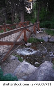 A vertical, high-angle shot capturing a rustic wooden footbridge spanning across a shallow, rocky mountain stream. The bridge features cross-braced wooden railings with a metal mesh lining, leading into a dense forest of tall pine trees. Below the bridge, clear water flows rapidly over dark stones and boulders, creating white ripples in the current. The scene is captured in soft, natural daylight, emphasizing the peaceful, wilderness setting.