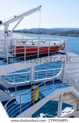 A vertical high-angle shot showcasing a red and white rescue lifeboat on a crane deck above blue painted stairs with a bright yellow trash bin on a passenger car ferry moving on deep blue sea water along the coast of Aegina Greece during a sunny summer