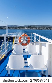 A vertical high-angle shot showcasing white plastic passenger seats and an orange lifebuoy on a blue painted deck of a ferry leaving a foaming white wake trace on deep blue sea water moving away from the coastal town and hills of Aegina Greece under a clear summer sky