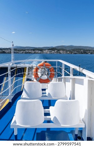 A vertical high-angle shot showcasing white plastic passenger seats and an orange lifebuoy on a blue painted deck of a ferry leaving a foaming white wake trace on deep blue sea water moving away from the coastal town and hills of Aegina Greece under a clear summer sky