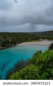 Vertical high-angle view of a beautiful turquoise sea cove and sandy beach under a stormy overcast sky in Menorca
