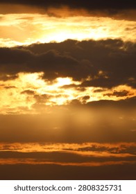 A vertical, high-contrast photograph capturing a dramatic and vibrant sunset breaking through a heavy layer of clouds.

Key Elements:
The Sky and Lighting: The scene is dominated by intense shades of gold, orange, and amber. The sun is partially obscured, creating a brilliant, glowing focal point near the upper-middle section of the image that radiates light outward.

The Clouds: Thick, dark gray and black clouds stretch horizontally across the frame, heavily contrasting against the fiery background. Smaller, fractured cloud formations gather directly around the sun, catching the bright light along their edges.

Crepuscular Rays: Faint sunbeams or light rays filter downward through gaps in the cloud cover, illuminating the hazy, golden atmosphere below.

The image has a powerful, almost apocalyptic or ethereal quality, emphasizing the stark contrast between the dark storm clouds and the brilliant light of the setting sun.
