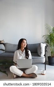 Vertical Image Of Ambitious Freelance Girl Sitting At Home With Laptop And Looking Outside Window, Smiling. Woman Working On Computer In Living Room.