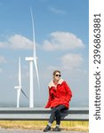 Vertical image of Caucasian woman with red coat sit on barrier of roadside and use mobile phone on mountain near windmill or wind turbine also look to right side.