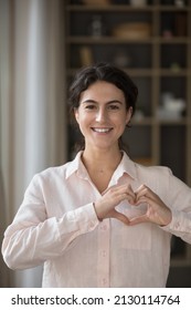 Vertical image cheerful attractive young Hispanic woman making heart symbol with fingers near chest, showing gratitude feeling peaceful. Happy kind female volunteer expressing support, charity concept