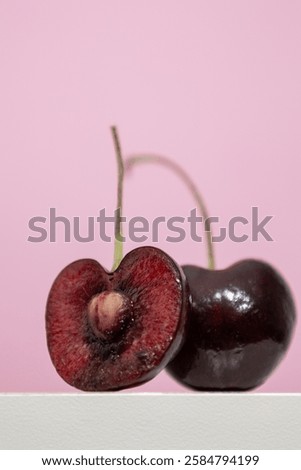 Vertical image of a cherry cut in half on a white wooden shelf on a pink background, small round red fruit with a seed in the center