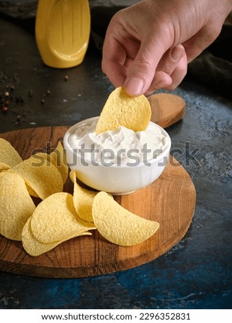 Vertical image close-up of a man dips potato chips into sauce in a white bowl on a wooden cutting board. On a dark blue abstract background.
